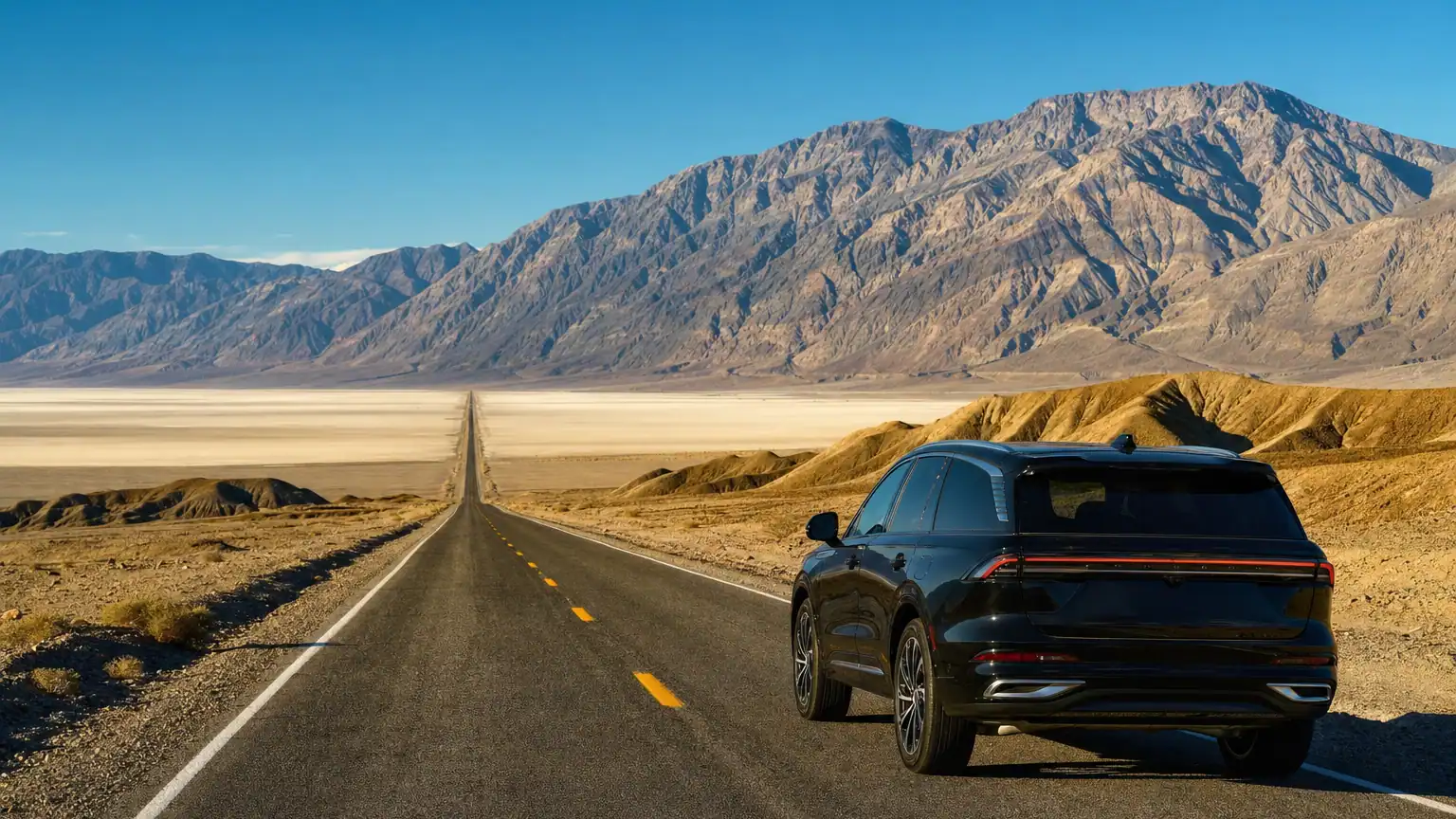 Black luxury SUV on an open desert highway through Death Valley with salt flats and mountains at golden hour